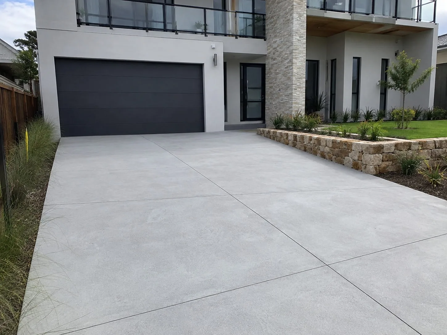 Broad plain concrete driveway with saw-cut panels leading to a dark double garage at a rendered Newcastle home